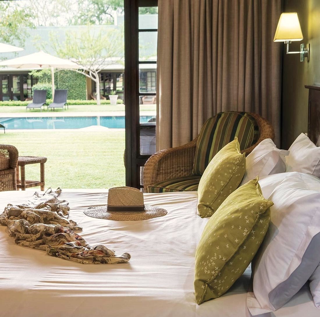 A quiet lodge bedroom opening onto a garden and pool, with a straw hat resting on the bed